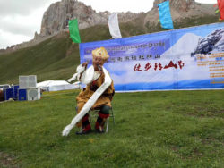 A Gesar bard performs in Yushu Tibetan Autonomous Prefecture in front of a sign with Chinese writing on it, and mountains in the background.